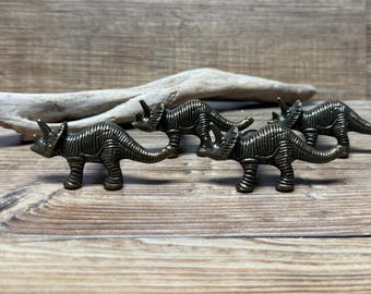 May include: Four bronze-colored dinosaur-shaped cabinet knobs. The dinosaurs are in a line, facing the same direction. The dinosaurs are detailed with textured skin and horns.
