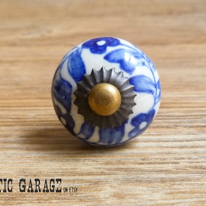 Può includere: Pomello rotondo in ceramica con base bianca e motivo floreale blu. Il pomello ha un centro color bronzo e una base in metallo scuro. L'immagine è su una superficie di legno.