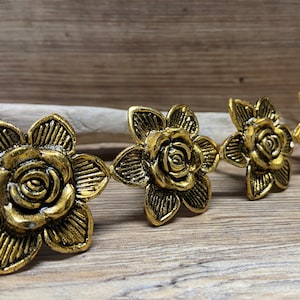 May include: Four gold-toned metal drawer knobs shaped like blooming flowers with textured petals. Each knob features a central rose-like bud surrounded by six radiating petals with etched lines, arranged in a row from largest to smallest.