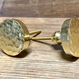 May include: Two gold-colored cabinet knobs with a hammered texture. The knobs have a round, flat face and a threaded post for mounting. The hardware is made of metal and is designed for use on drawers or cabinets. The knobs are displayed on a wooden surface.