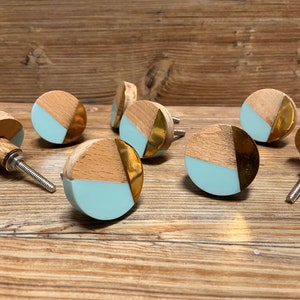 May include: A collection of wooden cabinet knobs with a light blue, gold, and natural wood design. The knobs are round with a metal screw for installation. The knobs are arranged on a wooden surface.
