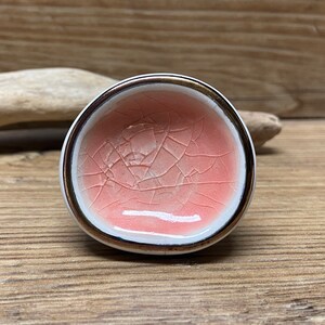 May include: A small, round ceramic bowl with a crackle glaze in shades of pink and white. The rim is trimmed with a gold band. The bowl is set against a wooden background.