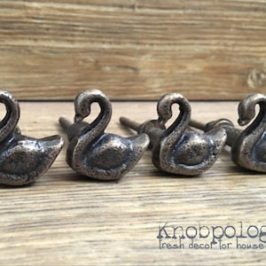 May include: Four antique-style swan-shaped cabinet knobs with a brushed metal finish. The knobs have a decorative screw detail and are designed for use on furniture.