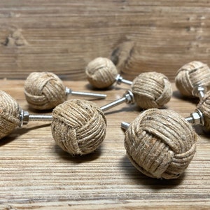 May include: Ten decorative cabinet knobs, each with a spherical design wrapped in natural twine. They are attached to a metal screw and nut. The knobs are displayed on a wooden surface.
