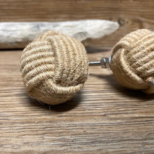 Peut inclure: Deux boutons de meuble décoratifs. Chaque bouton est une sphère faite de corde de jute naturelle étroitement enroulée, ressemblant à un nœud nautique. Les boutons sont fixés à une vis et un écrou métalliques. Le fond est une surface en bois.