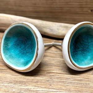 May include: Pair of white ceramic cabinet knobs with a turquoise interior and gold trim. The knobs are connected by a silver metal rod. The knobs are on a wooden surface.