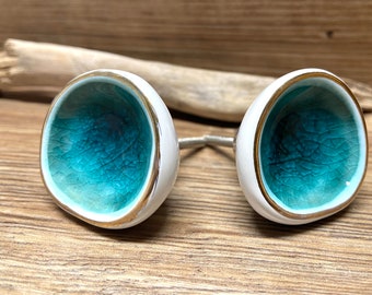 May include: Pair of white ceramic cabinet knobs with a turquoise interior and gold trim. The knobs are connected by a silver metal rod. The knobs are on a wooden surface.
