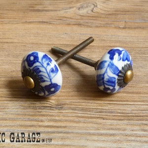May include: Two ceramic cabinet knobs with a white base and blue floral design. Each knob has a brass-colored center and a dark metal base. The knobs are attached to threaded rods, perfect for furniture upgrades.