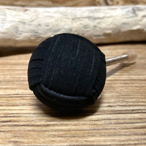 Può includere: Un tappo per bottiglia nero e rotondo con un design in tessuto strutturato. Il tappo ha uno stelo argentato e una punta arrotondata trasparente. Lo sfondo è una superficie di legno.