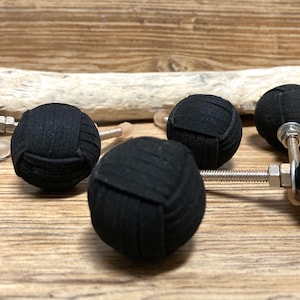 Può includere: Primo piano di diverse manopole per mobili a sfera in tessuto nero con ferramenta argentata. Le manopole sono disposte su una superficie di legno marrone chiaro, con un pezzo di legno alla deriva sullo sfondo. Le manopole hanno un aspetto strutturato e cucito.