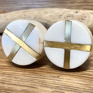 May include: Two white marble cabinet knobs with brass cross inlays. The knobs are round and sit on a wooden surface. The brass inlays create a decorative cross design on each knob.