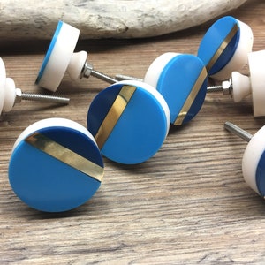 May include: A collection of decorative cabinet knobs. Each knob features a round design with a white base, a light blue and navy blue section, and a gold-colored stripe. The knobs have a screw for easy installation.
