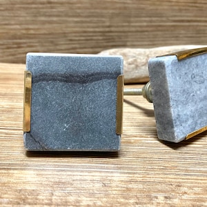 Könnte beinhalten: Zwei quadratische Schrankknöpfe. Jeder Knopf hat eine graue Marmorfläche mit goldfarbenen Metallakzenten. Die Knöpfe sind an einer silberfarbenen Metallschraube befestigt. Der Hintergrund ist eine hellbraune Holzoberfläche.