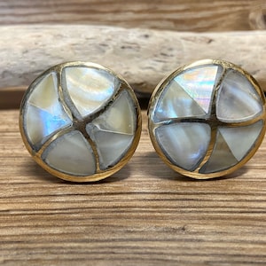 May include: A pair of round earrings with a gold-colored border. Each earring features a mosaic design of iridescent white and cream-colored pieces, creating a star-like pattern. The earrings are displayed on a wooden surface.