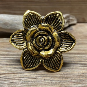 May include: A gold-toned metal ring featuring a six-petaled flower design. Each petal has a textured, striped pattern radiating from the centre, which is shaped like a small, layered rose. The metal has a dark, antiqued finish in the recessed areas.