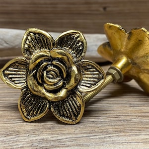 Könnte beinhalten: Zwei goldfarbene Metall-Möbelknöpfe in Form von sechsblättrigen Blumen mit einem zentralen Rosenmotiv. Jeder Knopf hat eine strukturierte Oberfläche und enthält einen Gewindebolzen aus Metall sowie eine kleine Mutter zur Befestigung an Möbeln.