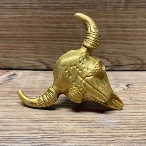May include: A gold-colored, decorative bull skull figurine. The sculpture has a textured surface with detailed horns and facial features. The bull skull is displayed on a wooden surface.