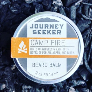 May include: A silver tin of beard balm with a black and orange label. The label reads "Journey Seeker Camp Fire" and "Hints of whiskey & rain, with notes of poplar, aspen, and birch." The label also reads "Beard Balm" and "2 oz 59.14 ml".