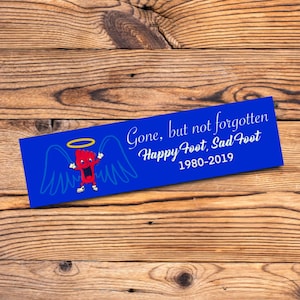 May include: A blue rectangular memorial plaque with the text "Gone, but not forgotten Happy Foot, Sad Foot 1980-2019." It features a cartoon foot with a halo and wings. The plaque is set against a wooden background.