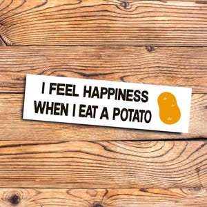 May include: A white sticker with the text "I FEEL HAPPINESS WHEN I EAT A POTATO" in black, alongside an orange potato graphic. The sticker is placed on a wooden surface.