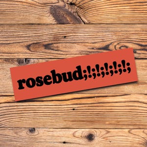 May include: An orange rectangular sticker with the word "rosebud" in black, followed by a series of exclamation points and semicolons. The sticker is placed on a wooden surface with visible wood grain.