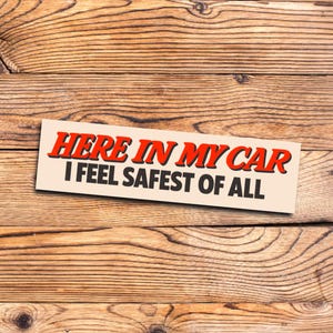 May include: A rectangular sticker with the text "HERE IN MY CAR I FEEL SAFEST OF ALL" in red and black lettering. The sticker is on a light beige background, set against a wooden plank backdrop.