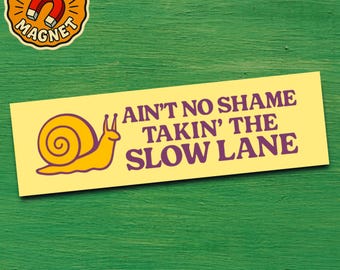 Op de afbeelding: Een gele bumpersticker met de tekst "AIN'T NO SHAME TAKIN' THE SLOW LANE" in paars. Een gele slakafbeelding staat links. Een ronde magneet met de woorden "BUMPER MAGNET" en een magneetafbeelding staat linksboven.