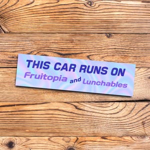 May include: A rectangular sticker with the text "THIS CAR RUNS ON Fruitopia and Lunchables" in blue and purple, set against a gradient background. The sticker is placed on a wooden surface with a natural wood grain.
