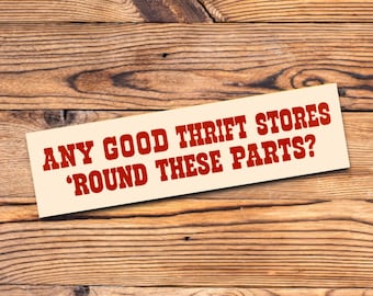 Op de afbeelding: Een rechthoekig bord met de tekst "ANY GOOD THRIFT STORES 'ROUND THESE PARTS?" in rode letters. Het bord staat op een houten achtergrond met zichtbare houtnerf.