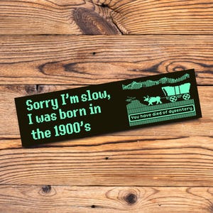 May include: A black rectangular sticker with the text "Sorry I'm slow, I was born in the 1900's" in a pixelated font. The right side features a pixelated image of a covered wagon and oxen. Below the image is the text "You have died of dysentery."