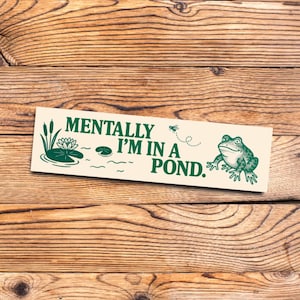 Puede incluir: Un letrero rectangular crema con el texto "MENTALLY I'M IN A POND." presenta una rana verde, nenúfares y juncos. El letrero tiene texto e ilustraciones verdes, sobre un fondo de madera.