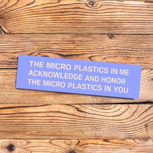 May include: A light blue rectangular sign with white text that reads: "THE MICRO PLASTICS IN ME ACKNOWLEDGE AND HONOR THE MICRO PLASTICS IN YOU." The sign is set against a wooden background.