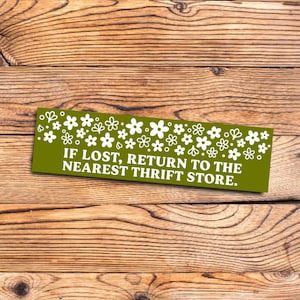 May include: A rectangular green sign with white floral designs and the text "IF LOST, RETURN TO THE NEAREST THRIFT STORE." The sign is set against a wooden background with a natural wood grain.