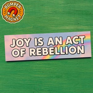 May include: A bumper magnet and a rectangular sticker with the text "JOY IS AN ACT OF REBELLION". The sticker has a pastel rainbow and sky background. The bumper magnet has a magnet graphic.
