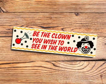 May include: A yellow and black banner with a red clown face and a red horn. The banner reads "Be the clown you wish to see in the world."