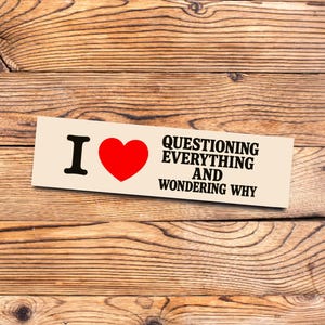 Puede incluir: Una pegatina rectangular beige con el texto "I ❤️ QUESTIONING EVERYTHING AND WONDERING WHY". La "I" es negra, el corazón es rojo y el texto es negro. La pegatina está sobre una superficie de madera.