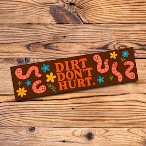 May include: A brown rectangular sticker with the text "DIRT DON'T HURT" in orange. The sticker features cartoon worms, blue and yellow flowers, and green leaves. The sticker is on a wooden surface.