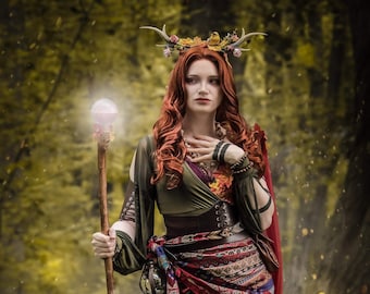 May include: A woman in a forest setting, wearing a green dress, a brown corset, and a decorative belt with red trim. She has red hair, a floral and antler headpiece, and holds a staff with a glowing orb.