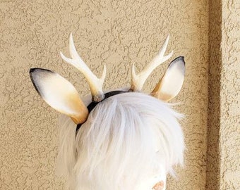 May include: A white wig with a deer antler headband. The headband is made of faux antlers and has a black band. The wig is styled in a short, choppy bob.