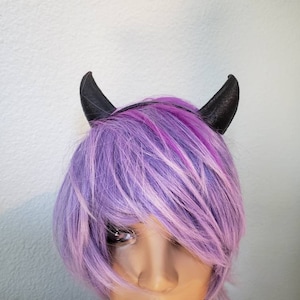 Può includere: Un cerchietto nero con due corna di diavolo. Il cerchietto è indossato da un manichino con capelli corti e viola.