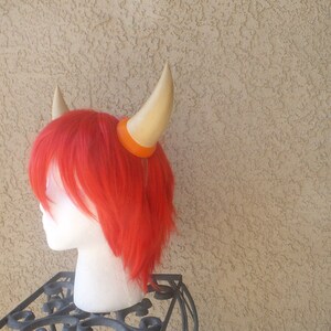 May include: A red wig with two gold horns attached to it. The horns are attached with orange elastic bands.
