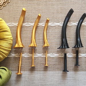 Può includere: Un set di sei gambe per mobili curve di varie dimensioni, composto da tre gambe color oro metallizzato e tre gambe nero opaco. Ogni gamba presenta una base svasata e include un bullone filettato in metallo abbinato per il fissaggio.