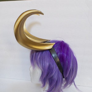 May include: A gold crescent moon-shaped headband with a black elastic band. The headband is worn on a purple wig.
