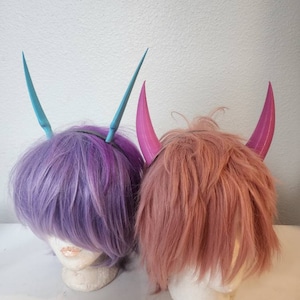 Peut inclure: Deux perruques avec des cornes de diable, une perruque est violette et l'autre est rose.