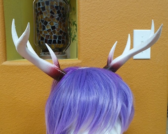 May include: A white wig with vibrant purple hair styled in a layered cut, featuring a pair of attached deer antlers. The antlers are primarily white with dark red and black shading at the base, mounted on a headband worn by a white mannequin head.