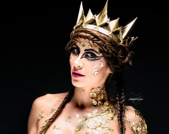 May include: A woman with gold leaf makeup and a gold crown, wearing a gold crown with a pointed design. Her hair is styled in two braids. The background is black.