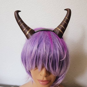 May include: A headband with two brown horns with gold accents. The headband is worn on a purple wig.