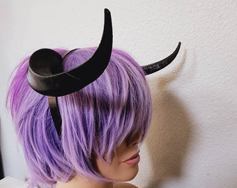 Könnte beinhalten: Ein schwarzes Stirnband mit zwei daran befestigten schwarzen Hörnern. Das Stirnband wird von einer Puppe mit lila Haaren getragen.