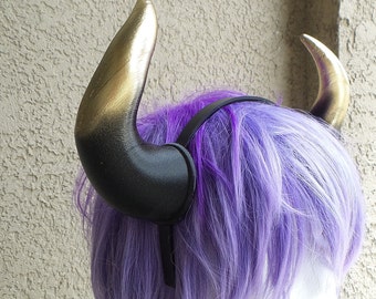 Könnte beinhalten: Ein Paar schwarze und goldene Hörner mit einem Farbverlauf. Die Hörner sind an einem schwarzen Stirnband befestigt. Das Stirnband wird auf einer lila Perücke getragen.