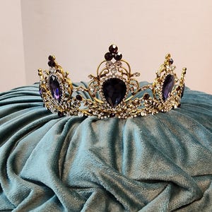 Puede incluir: Una corona dorada con diamantes de imitación morados y transparentes. La corona tiene un diseño en forma de corazón en el centro.
